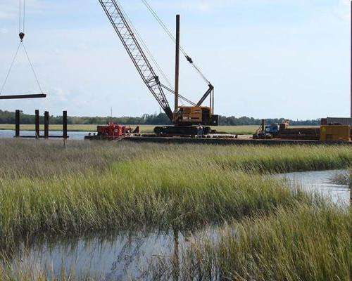 A crane lifts a beam across a swampy grassy marsh.