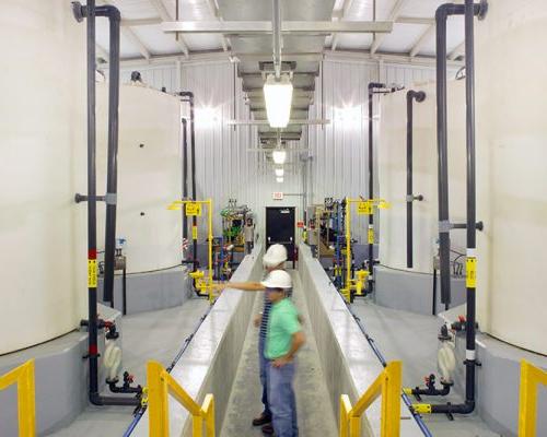 Interior photo of the facility with large white tanks. Men in hardhats checking their status.